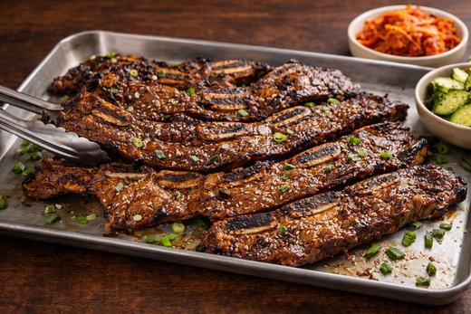 Korean BBQ beef short ribs (Galbi) grilled to perfection on a tray with sesame seeds, green onions, and side kimchi and cucumber salad.