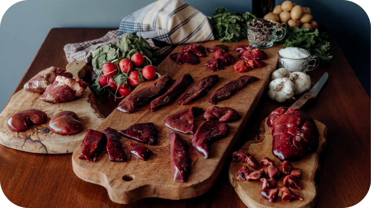 Assorted raw organ meats including liver, heart, and kidney arranged on wooden boards with fresh vegetables, garlic, and herbs for preparation.  Image source