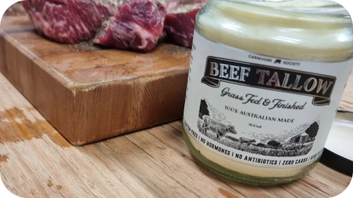 A jar of Carnivore Society Beef Tallow sits next to a wooden butcher block with several cubes of raw, seasoned marbled beef.