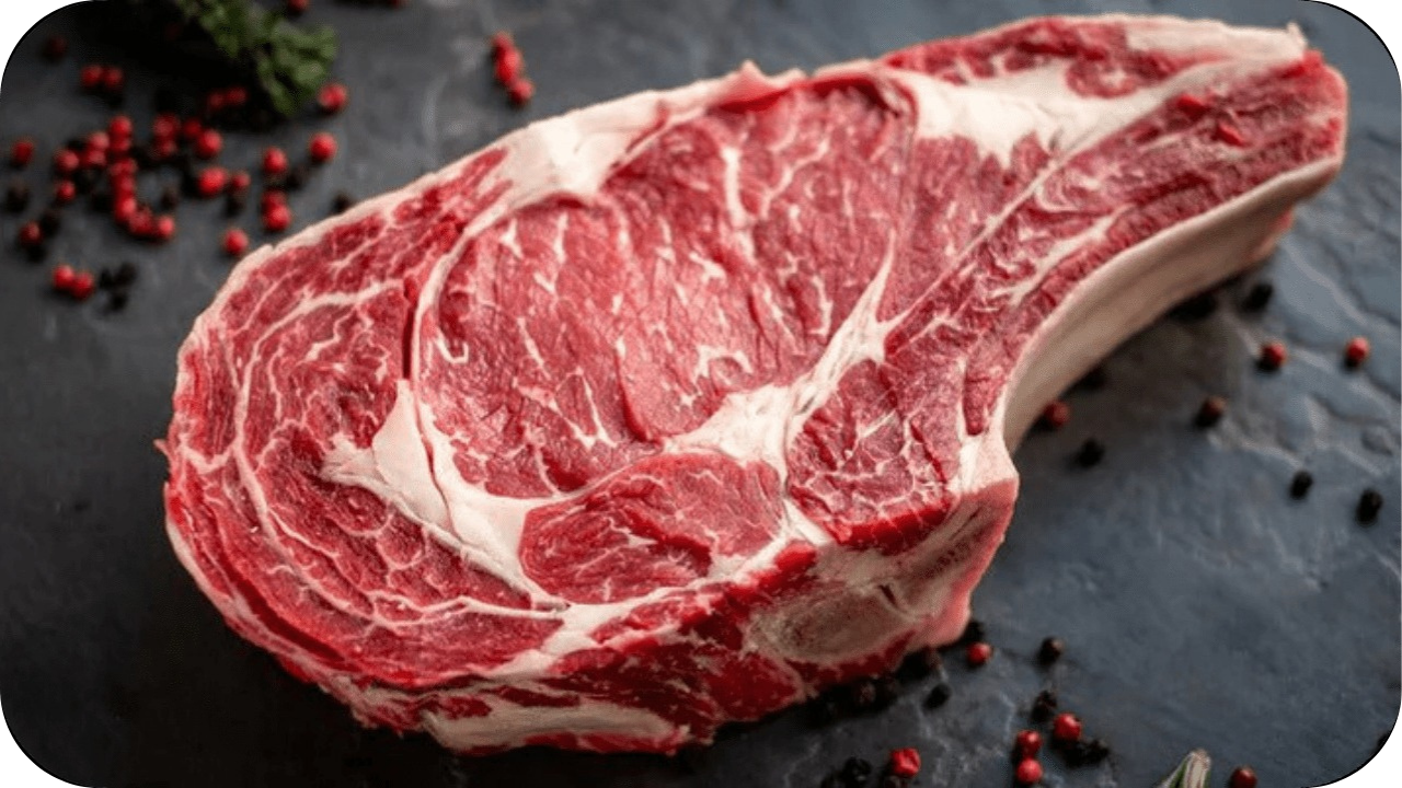 Raw ribeye steak on dark stone surface with scattered peppercorns and herbs