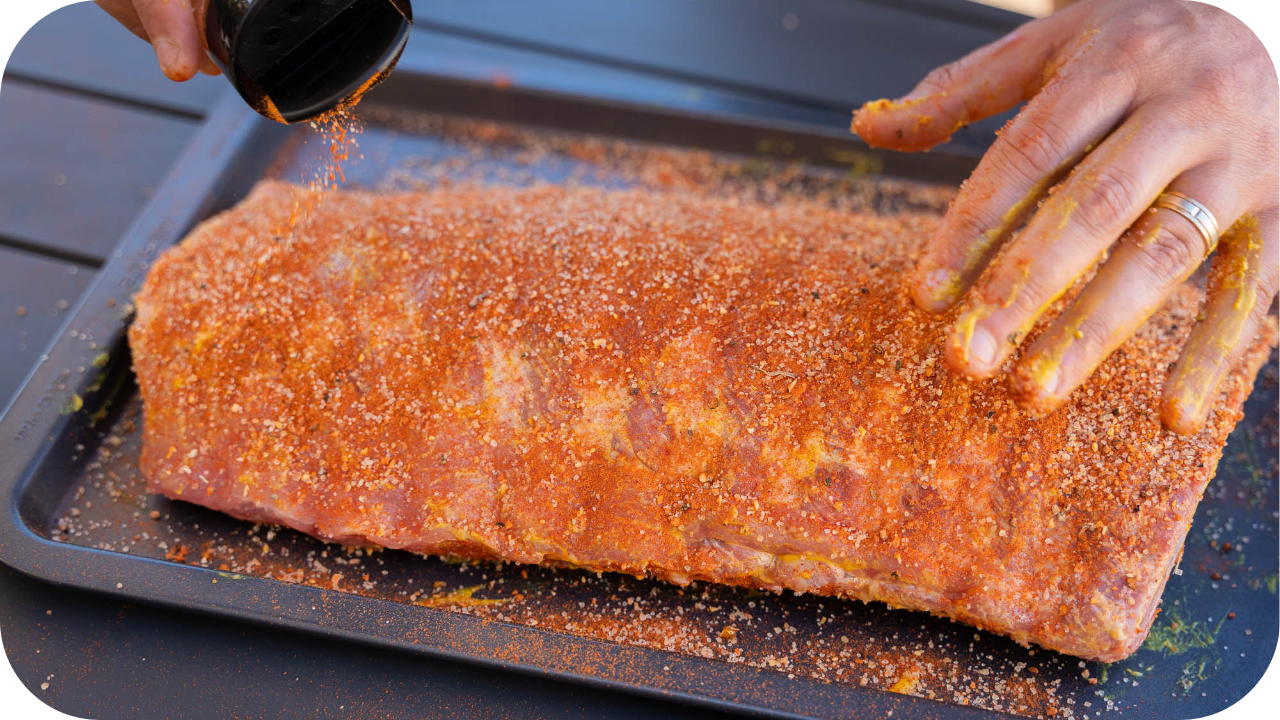 Hands seasoning a large rack of ribs with a dry spice rub on a baking tray, ready for grilling or smoking.