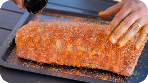 Hands seasoning a large rack of ribs with a dry spice rub on a baking tray, ready for grilling or smoking.