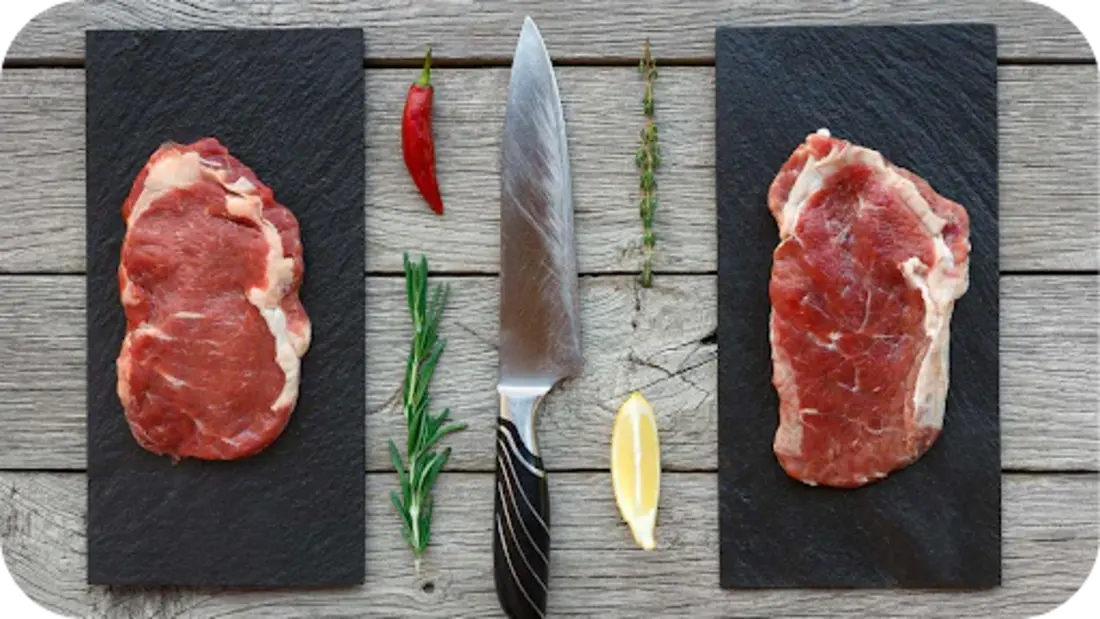 Two raw steaks displayed on black boards with herbs, chilli, lemon, and a knife on rustic wood, showing grass-fed vs grain-fed.