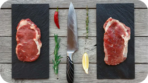 Two raw steaks displayed on black boards with herbs, chilli, lemon, and a knife on rustic wood, showing grass-fed vs grain-fed.