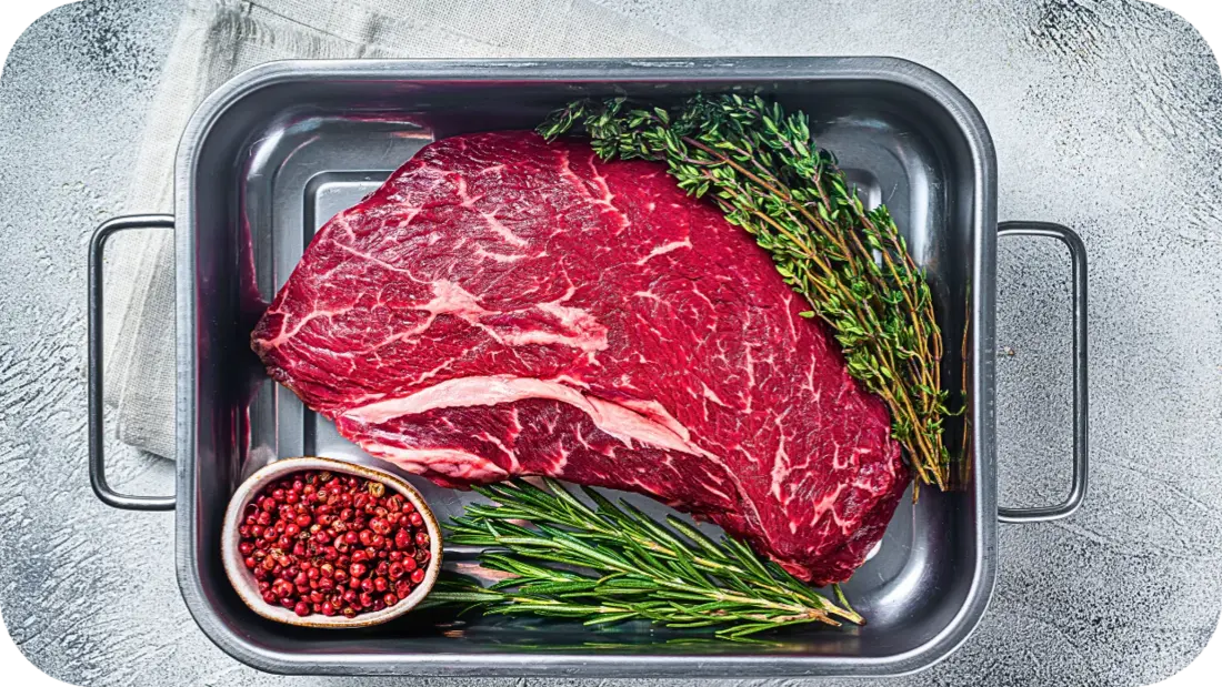 Fresh raw beef cut resting in a roasting pan with rosemary, thyme, and a bowl of red peppercorns, ready for cooking on a rustic kitchen surface.