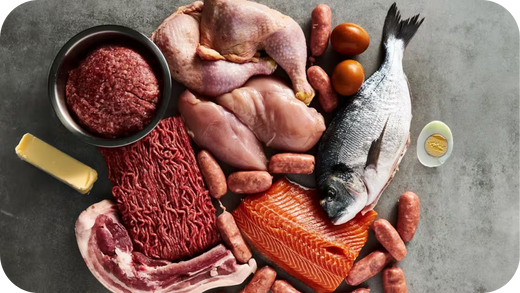 A variety of fresh meats and seafood including beef, chicken, salmon, sausages, and fish on a grey surface.