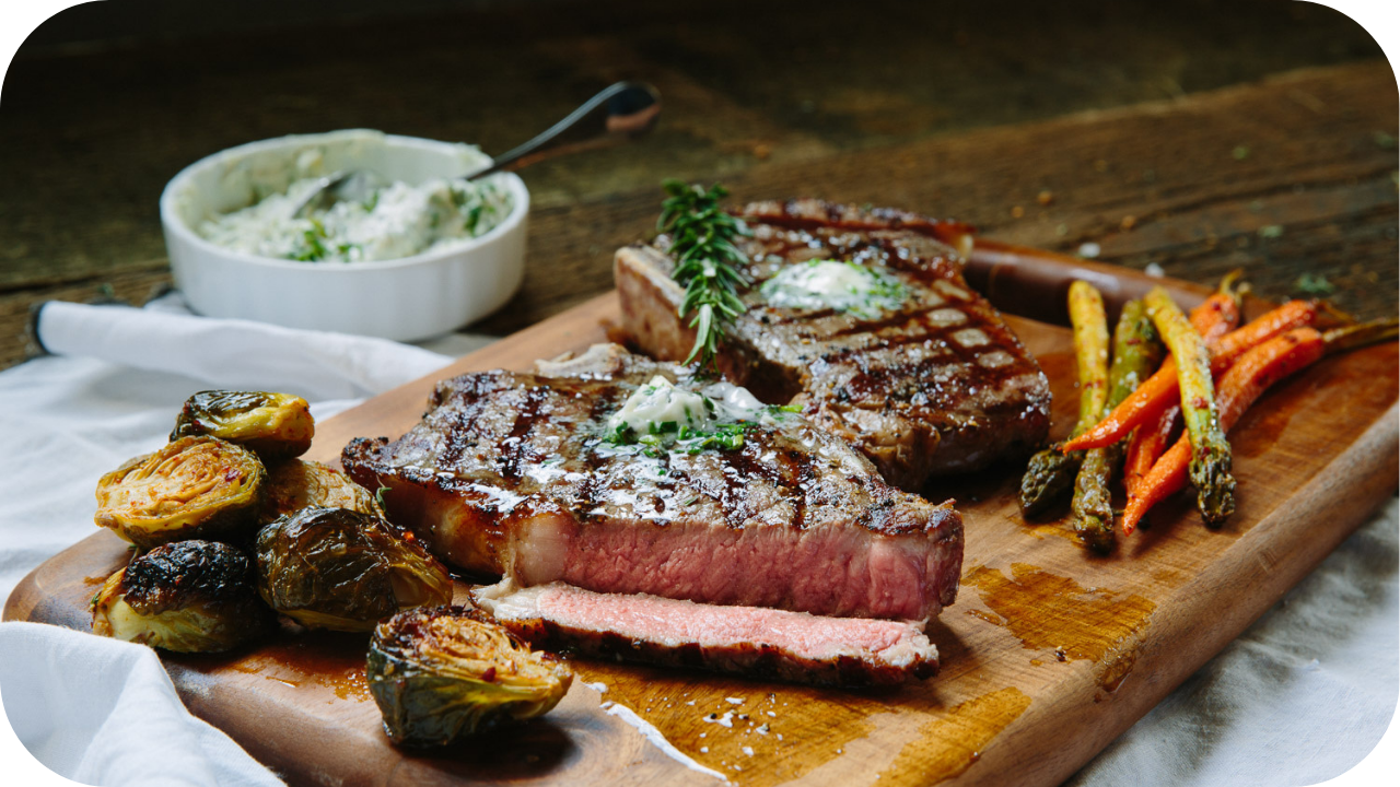 Grilled steak with herb butter served on a wooden board alongside roasted Brussels sprouts, carrots, and asparagus.