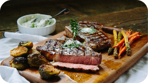 Grilled steak with herb butter served on a wooden board alongside roasted Brussels sprouts, carrots, and asparagus.
