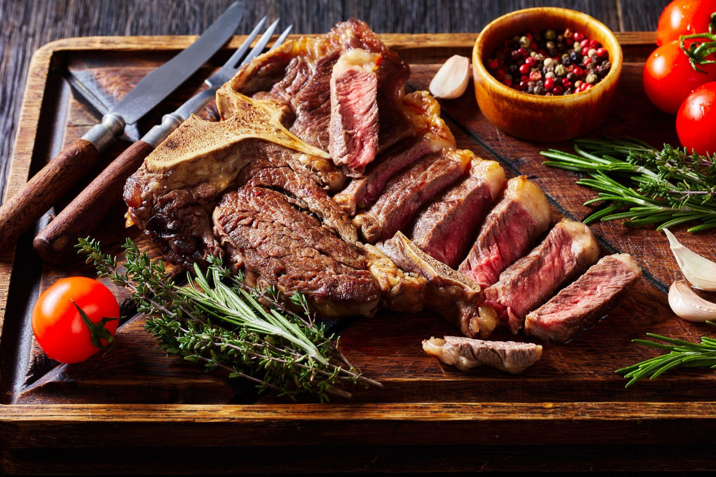 Sliced medium-rare Porterhouse steak with garlic tallow sear on a wooden board, garnished with fresh rosemary, thyme, cherry tomatoes, and garlic cloves