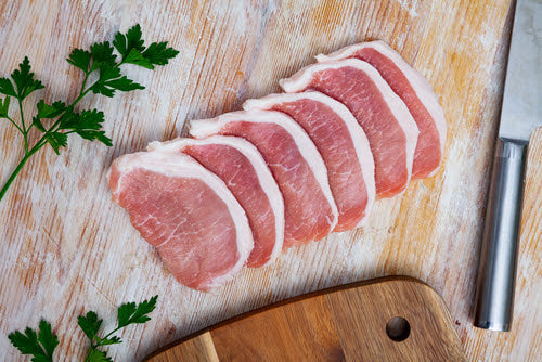 Six raw pork loin slices neatly arranged on a wooden surface, with fresh parsley, a chopping board, and a kitchen knife nearby.